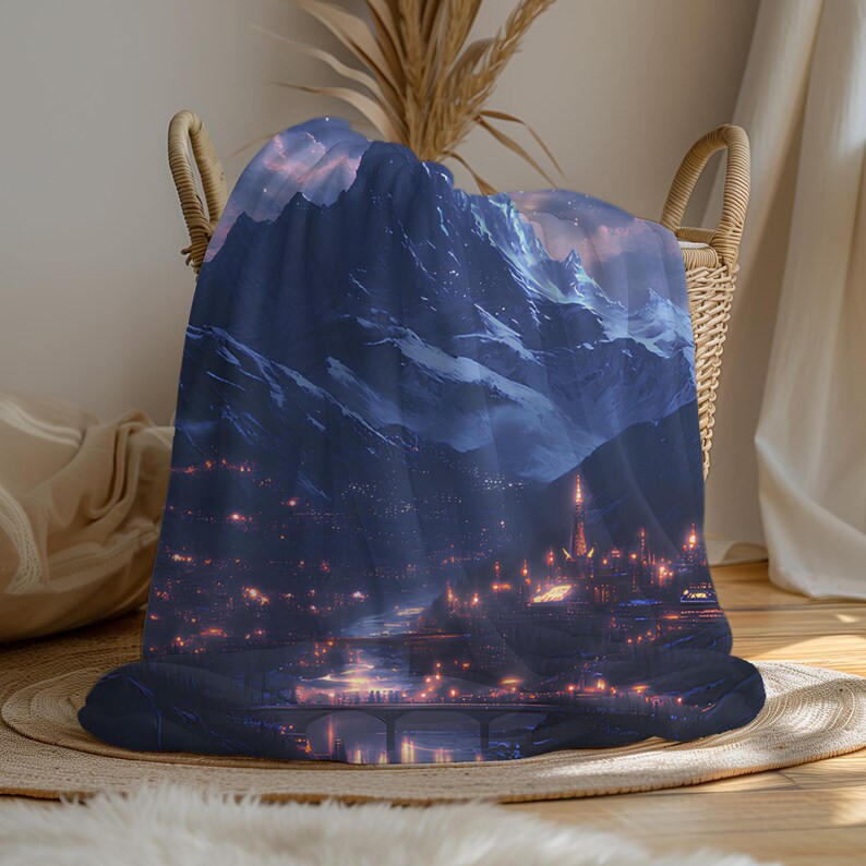 May include: A dark blue blanket featuring a mountain landscape with snow-capped peaks and a city at the base. The blanket has a detailed illustration of a city with glowing lights, a river, and a bridge.