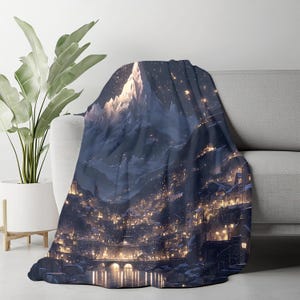 May include: A soft throw blanket with a detailed illustration of a mountain range and a city at night. The design features a snow-covered peak, a dark blue sky, and the warm glow of city lights. The blanket is draped over a grey sofa.