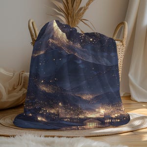 May include: A dark blue blanket with a detailed illustration of a mountain range at night. The design includes a city with illuminated buildings, a bridge, and a starry sky. The blanket is folded in a wicker basket.