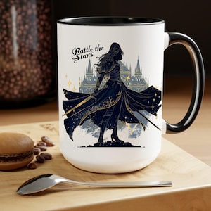 May include: White ceramic mug with a black interior and handle. The mug features a detailed illustration of a woman with a sword in front of a castle, with the text "Rattle the Stars". The design is in shades of blue and gold.
