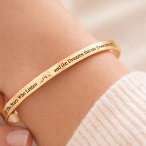 Puede incluir: Una pulsera de brazalete de color dorado con texto grabado. La pulsera se lleva en la muñeca, con el texto "To The Stars Who Listen and the Dreams that are Answered". La pulsera tiene un diseño sencillo y elegante.