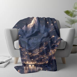 May include: A dark blue throw blanket draped over a white armchair. The blanket features a detailed illustration of a mountain range and a city at night, with warm yellow lights illuminating the scene. The design evokes a fantasy or landscape theme.