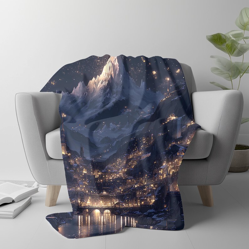 May include: A dark blue throw blanket draped over a white armchair. The blanket features a detailed illustration of a mountain range and a city at night, with warm yellow lights illuminating the scene. The design evokes a fantasy or landscape theme.