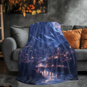 May include: A blue throw blanket featuring a detailed illustration of a mountain range and a city at night. The artwork depicts a river, bridge, and buildings with illuminated windows. The blanket is draped over a gray sofa with orange pillows.
