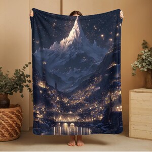 May include: A dark blue blanket with an illustration of a mountain range and a city at night. The artwork shows a starry sky, illuminated buildings, and a bridge over a river. The blanket is held up by a person.