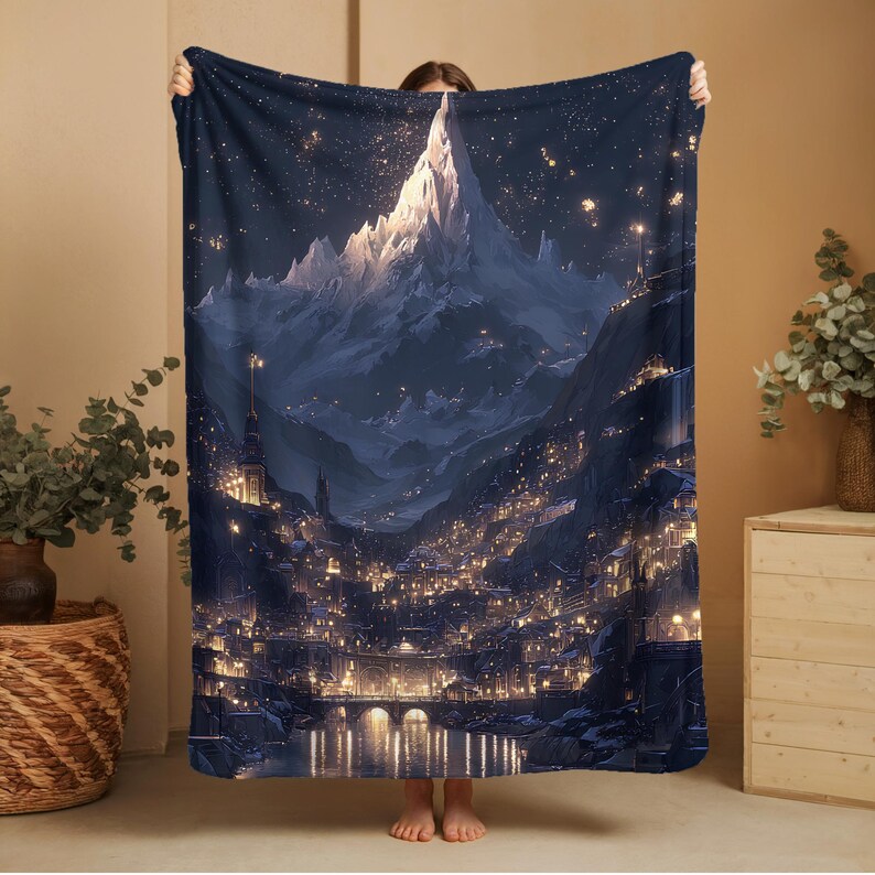 May include: A dark blue blanket with an illustration of a mountain range and a city at night. The artwork shows a starry sky, illuminated buildings, and a bridge over a river. The blanket is held up by a person.