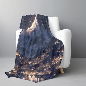 May include: A dark blue blanket with a mountain and town illustration draped over a white armchair. The blanket depicts a nighttime scene with a mountain range and a town illuminated by many lights. The design creates a cosy, nighttime atmosphere.