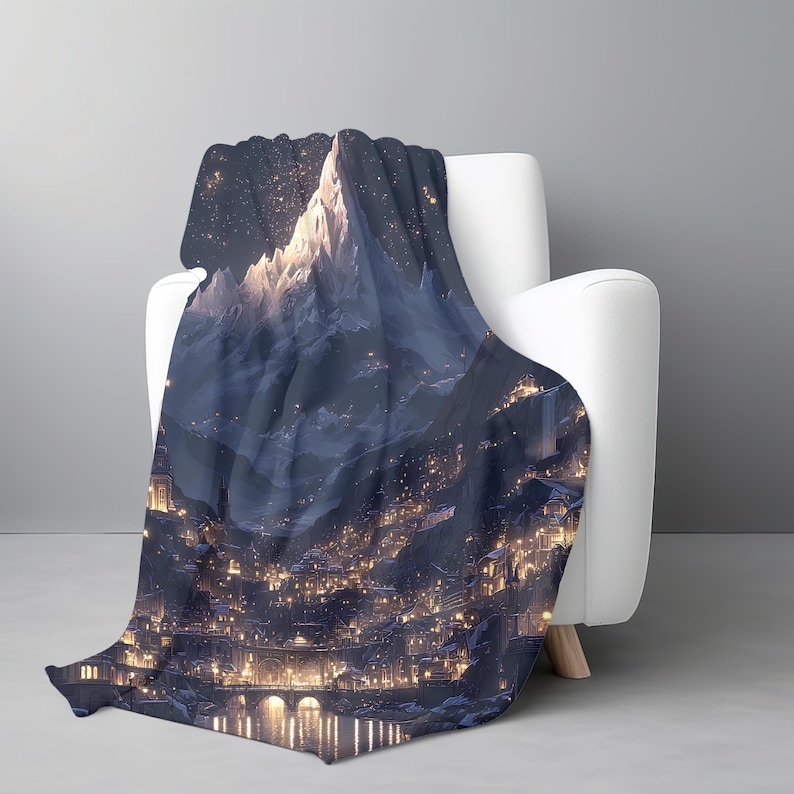 May include: A dark blue blanket with a mountain and town illustration draped over a white armchair. The blanket depicts a nighttime scene with a mountain range and a town illuminated by many lights. The design creates a cosy, nighttime atmosphere.