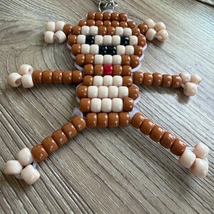 May include: A handmade monkey keychain crafted from brown, tan, black, white, and red beads. The monkey has a smiling face, outstretched arms, and legs. The keychain is attached to a silver-colored clasp.