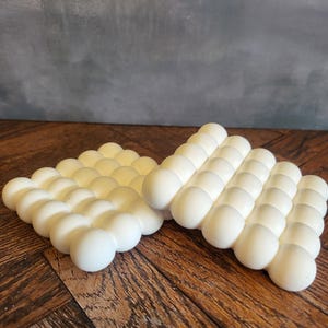 May include: Two off-white, bubble-shaped trivets are displayed on a wooden surface. The trivets are made of a soft material and are arranged side by side. The background is a muted grey wall.