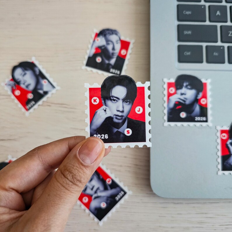 May include: A collection of small, square stickers resembling postage stamps. Each sticker features a black and white portrait against a red background, with the year "2026" printed on them. The stickers are scattered on a light-colored surface near a laptop.