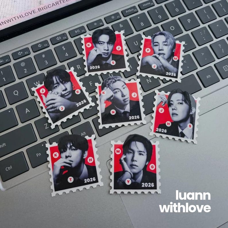 May include: A collection of seven postage stamp-style stickers featuring portraits of individuals. Each sticker has a red background with a black and white image, the year "2026", and a letter. The stickers are arranged on a laptop keyboard. The text "luann withlove" is visible.