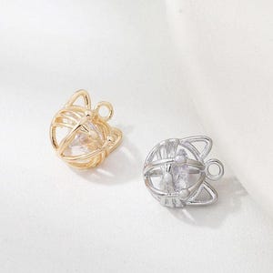 Könnte beinhalten: Zwei katzenförmige Anhänger. Einer ist goldfarben, der andere silberfarben. Jeder Anhänger hat ein käfigartiges Design mit einem klaren, facettierten Stein im Inneren. Die Anhänger haben eine kleine Schlaufe zum Befestigen an Schmuck.