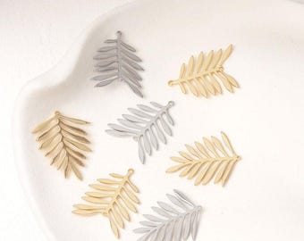 May include: An assortment of metal leaf charms in gold and silver tones. The charms are shaped like stylized palm leaves, each with a small loop for attaching to jewelry or other crafts. The leaves are arranged on a white surface.