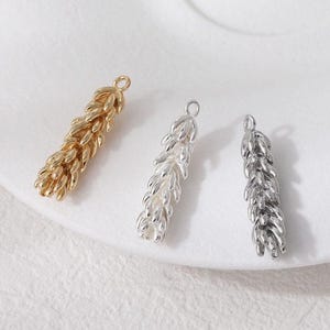Könnte beinhalten: Drei metallene Weizenähren-Anhänger in Gold, Silber und einem dunkleren Silberton. Jeder Anhänger hat oben eine kleine Schlaufe zum Befestigen an Schmuck. Die Anhänger sind auf einer weißen Oberfläche angeordnet.