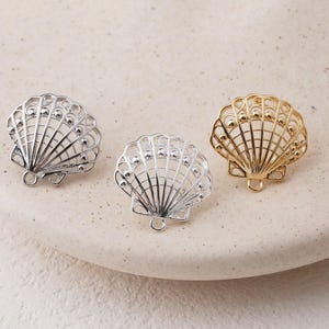 May include: Three decorative seashell charms in silver and gold tones. Each charm has an openwork design with a small loop at the bottom for attaching to jewellery or other crafts. The charms are arranged on a neutral surface.