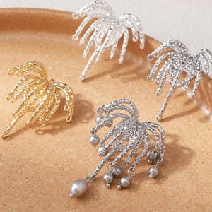 May include: Four decorative earrings in gold and silver tones, arranged on a tan surface. The earrings have a textured, abstract design, resembling a stylized palm frond. One silver earring features dangling pearl accents.