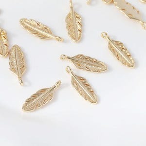 May include: Gold-coloured feather charms with detailed veining. These small, decorative pendants are designed for jewellery making or craft projects. The charms are arranged on a white surface.
