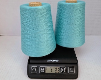 May include: Two turquoise yarn cones are on a black digital scale. The scale shows a weight of 1.23 kg. The yarn cones are cylindrical with a wooden core at the top. The scale has a power button, a tare button, and a hold button.