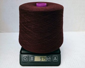 May include: A large cone of dark maroon yarn with a purple plastic centre core. The yarn cone is positioned on a black digital scale that displays a weight of 979 grams. The scale features buttons for power, tare, and hold functions.