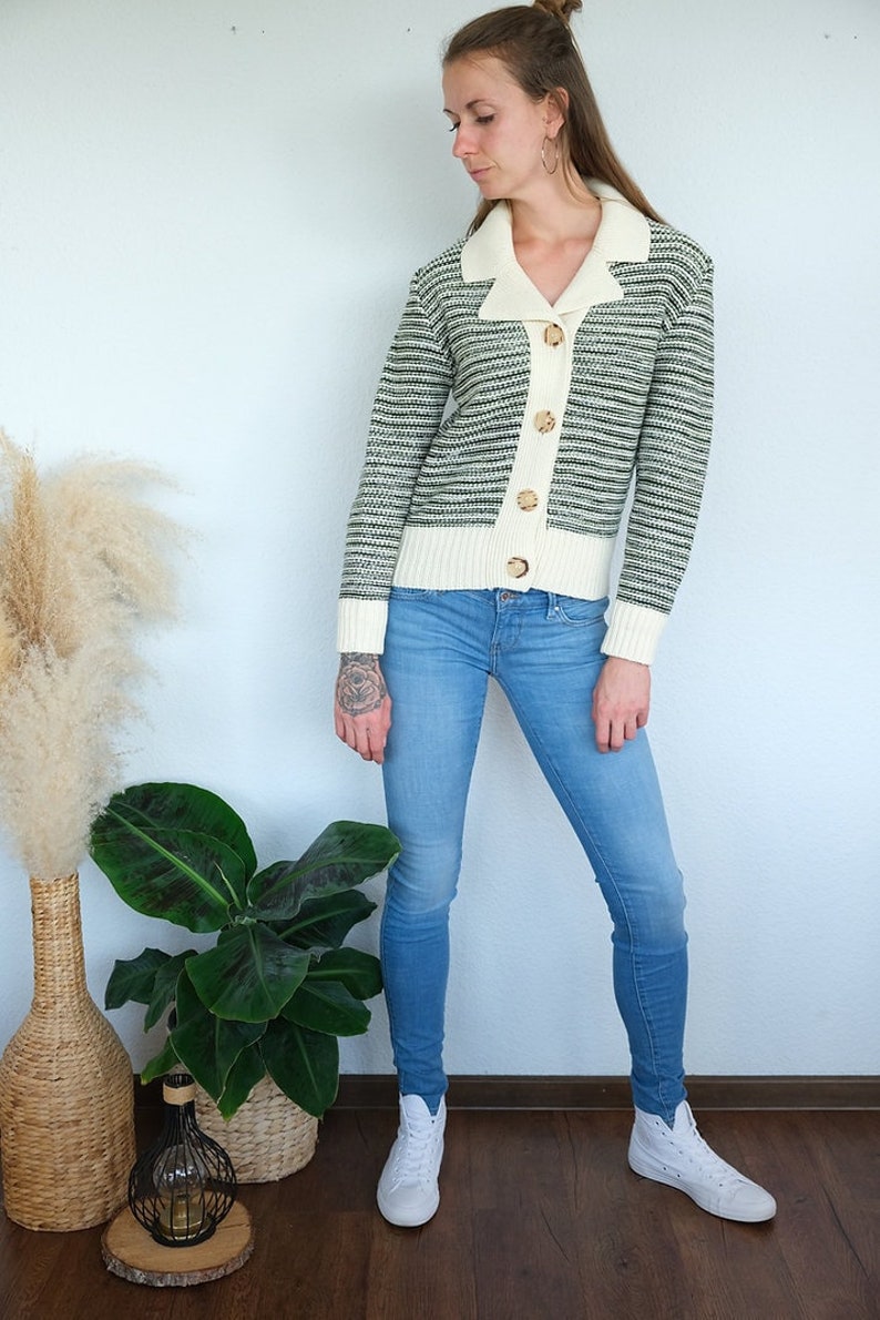 Weibliches Model trägt Strickjacke in creme,Bündchen und Kragen in creme. Rest grün gestreift. Dazu trägt sie eine hellblaue Jeans und weiße Sneaker.