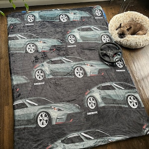 May include: A black blanket featuring a repeating pattern of gray sports cars with the text "TUNERHYPE". A black steering wheel is on the blanket. A dog bed with a dog is in the background.