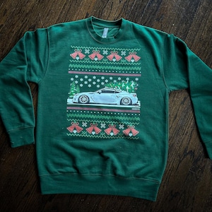 Peut inclure: Sweat-shirt vert avec un motif festif. Le graphisme présente une voiture blanche avec un sapin de Noël sur le dessus, entourée de flocons de neige, de cloches et d'un motif répétitif. Le texte "FINE TYPE" est visible.