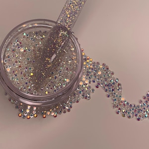 Peut inclure: Un pot transparent rempli de strass blancs irisés, avec une petite quantité de strass éparpillés sur une surface blanche. Un flacon de vernis à ongles transparent avec des paillettes irisées est présenté sur l'image.