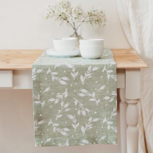 Pode incluir: Um caminho de mesa verde sálvia com um padrão floral branco, exibido sobre uma mesa de madeira. O caminho de mesa apresenta um design repetido de flores brancas estilizadas e pontos. Tigelas e pratos brancos empilhados na mesa, com um vaso de flores brancas ao fundo.