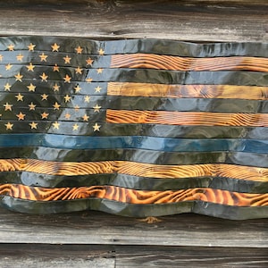 May include: A wall-mounted wooden American flag with a wavy, three-dimensional design. The flag features alternating charred black and natural wood grain stripes, with a single horizontal blue stripe and carved stars on a black field.