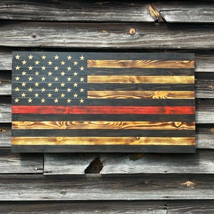 May include: A wooden American flag with a thin red line, featuring a distressed finish. The flag's stars are gold, and the stripes alternate between black and a lighter wood tone. The flag is mounted on a rustic wooden background.