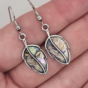 May include: A pair of leaf-shaped earrings with iridescent abalone shell inlays. The earrings have a silver-colored metal frame and hook closures. The abalone shell displays a range of colors, including pink, green, and gold, creating a natural, eye-catching design.