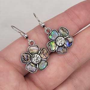 May include: A pair of silver dangle earrings with a flower design. The flower is made of iridescent abalone shell and has a clear rhinestone in the center.