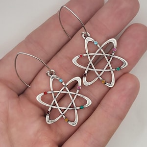 May include: Silver-toned atom earrings with colorful gemstone accents. Each earring features a stylized atom design with a central nucleus and orbiting electrons represented by small, multi-colored gems. The earrings have a hook closure.