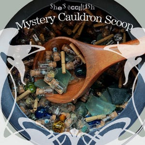 May include: A black cauldron filled with small glass bottles, cork stoppers, rolled paper scrolls, and green sea glass, with a wooden scoop resting on top. The text "She's occultish Mystery Cauldron Scoop" is printed in white across the top.