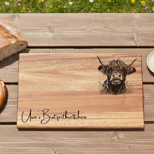 Könnte beinhalten: Holzschneidebrett mit einer Illustration einer Hochlandkuh und dem Text "Uwe's Brotzeitbrettchen". Das Brett liegt auf einem Holztisch mit Brot, Brezel, Butter und Kaffee.
