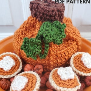 May include: A crocheted pumpkin with green leaves sits on a plate with crocheted pumpkin pie decorations and small brown and orange pumpkins.