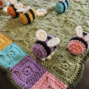 May include: A handmade crochet blanket featuring a green background with white cloud and flower accents. Adorned with several crochet bees in various colors, and a patchwork border in shades of yellow, teal, purple, and pink.