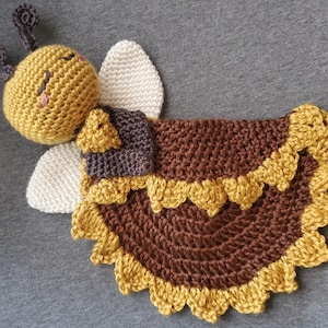 May include: A crocheted bee-shaped baby blanket with a brown and yellow sunflower design. The bee has a white and yellow striped body and a brown face.