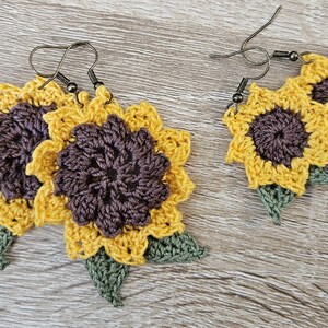 May include: A pair of crocheted sunflower earrings with green leaves. The earrings are yellow and brown with a dark brown centre.