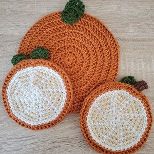 Puede incluir: Tres posavasos de ganchillo con forma de rodaja de naranja. El posavasos más grande es naranja con una hoja verde en la parte superior. Los dos posavasos más pequeños son naranja con un centro blanco y una hoja verde en la parte superior.