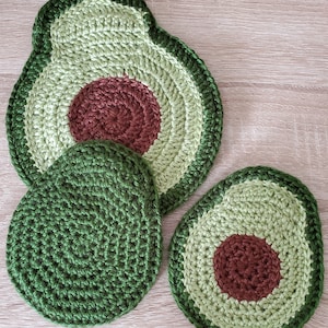 May include: Three handmade crochet avocado-shaped items. Two are avocado halves with a brown seed, and one is a solid green avocado pit. The items are in shades of green and brown, and are likely coasters or kitchen decor.