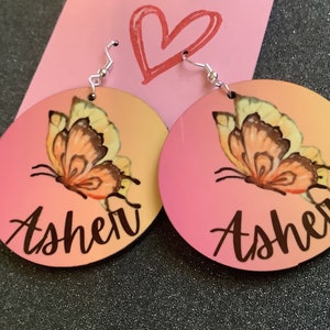 Op de afbeelding: Een paar ronde houten oorbellen in roze en oranje met een vlinderontwerp en de naam "Asher" in zwarte letters.