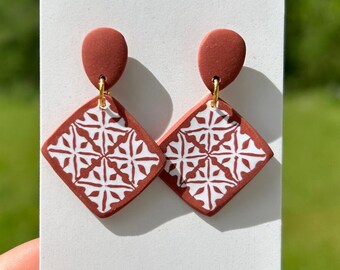 May include: A pair of terracotta-colored dangle earrings with a white geometric pattern. The earrings are displayed on a white card.