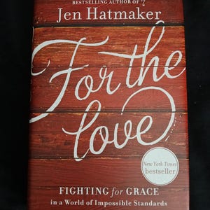 May include: Book cover with a red woodgrain background and white text that reads "For the love" in a cursive font. The title of the book is "Fighting for Grace in a World of Impossible Standards" and the author is Jen Hatmaker. The book is a New York Times bestseller.