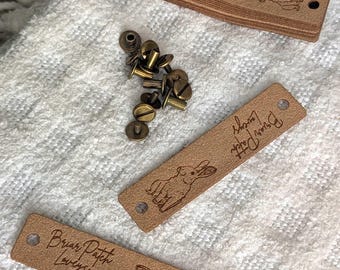 Op de afbeelding: Verschillende rechthoekige, lichtbruine leren labels met afgeronde hoeken, elk met de tekst "Briar Patch Lovelies" en een konijnenillustratie. Kleine messing klinknagels liggen in de buurt verspreid. De labels hebben voorgeboorde gaten.