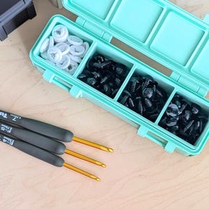 May include: A mint green plastic storage container with three compartments filled with white and black plastic safety eyes. Three crochet hooks with gold-colored metal tips and grey handles are positioned next to the container.