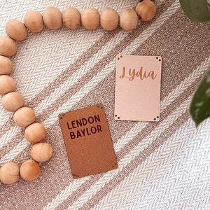 May include: Two personalized leather name tags. One tag is brown with the name "Lendon Baylor" and the other is pink with the name "Lydia".