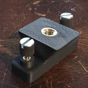 May include: A black, rectangular camera accessory with a brass-colored threaded insert and two silver-topped knobs. The knobs have a ridged texture. The accessory is set against a brown, textured surface.