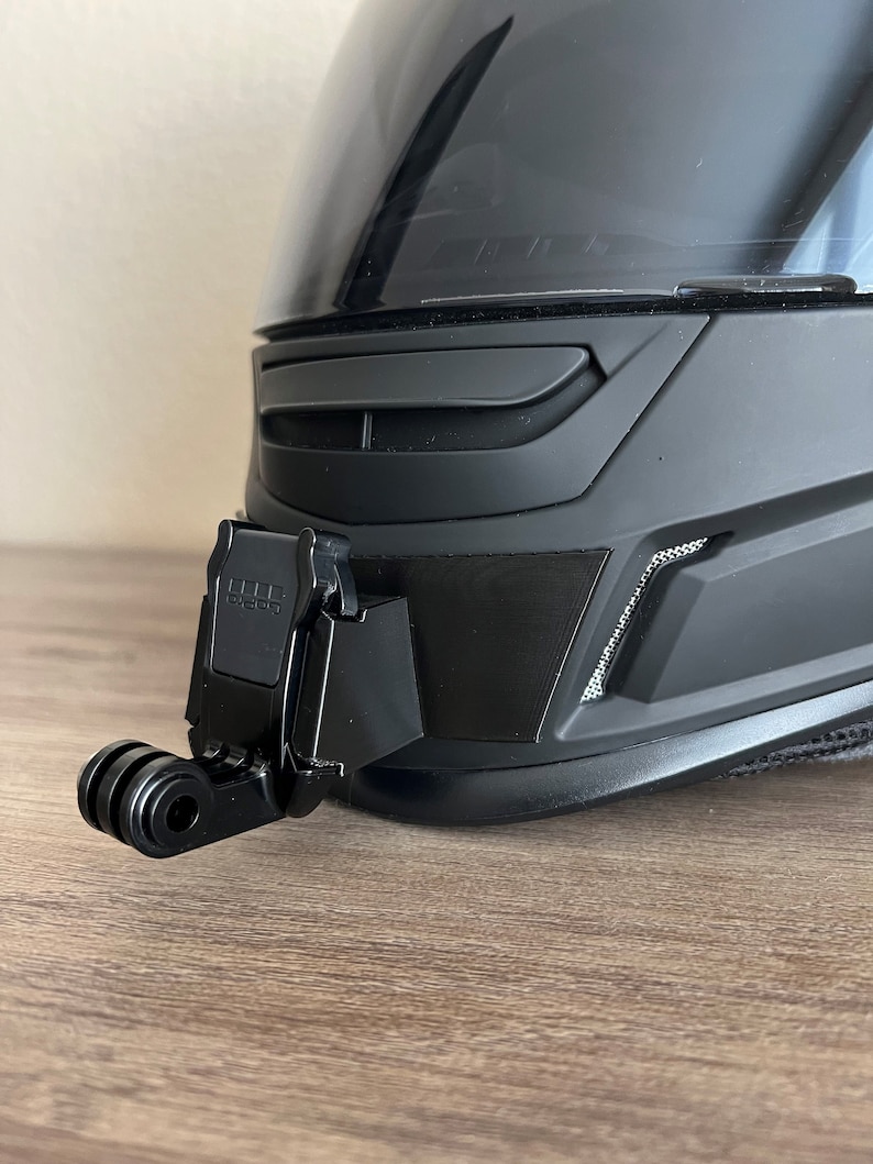 May include: Black motorcycle helmet with a GoPro mount attached to the front. The mount is a black plastic bracket with a GoPro camera attached to it.